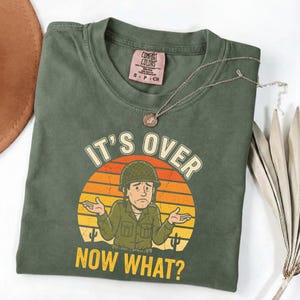 Puede incluir: Camiseta verde oliva con la frase "IT'S OVER NOW WHAT?" y un dibujo de un soldado de dibujos animados. El diseño presenta un atardecer en tonos naranja, amarillo y rojo. Un collar plateado con un colgante está sobre la camiseta doblada.