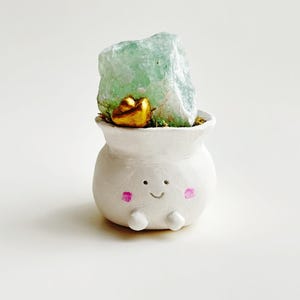 May include: A white ceramic planter with a smiling face and pink cheeks, holding a green crystal and a gold accent.