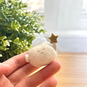 May include: A small white ceramic figurine of a cloud with a smiling face holding a gold star. The cloud has a white wing on one side.