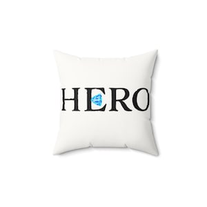 Puede incluir: Cojín cuadrado blanco con la palabra "HERO" impresa en negro. La letra "O" de "HERO" se reemplaza con un gráfico en forma de diamante azul y blanco. Artículo decorativo.