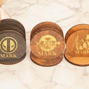 May include: A set of three wooden coasters with gold-colored designs and the name "MARK". One coaster features a Deadpool logo, another the X-Men logo, and the third a character silhouette.