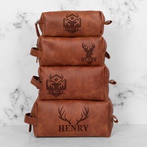 May include: Four stacked brown leather toiletry bags with engraved deer designs and the name "HENRY". The bags have a rectangular shape with a zipper closure and a handle on the side. The bags are in varying sizes.