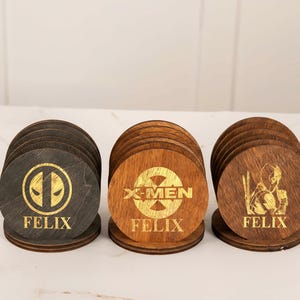 May include: Set of three wooden coasters with superhero logos and the name "FELIX" engraved in gold. The coasters feature designs from Deadpool, X-Men, and a third character. Each coaster is round and rests on a matching circular base.