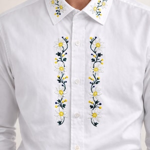 Puede incluir: Camisa blanca con bordado floral. El cuello y los paneles frontales presentan flores blancas bordadas con centros amarillos y hojas verdes. La camisa tiene un cuello clásico y mangas largas.