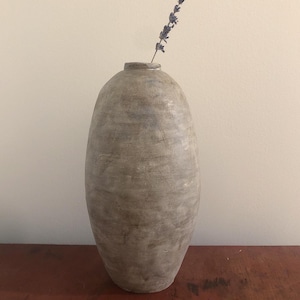 Puede incluir: Un jarrón de cerámica gris con forma ovalada redondeada. Un solo tallo de lavanda seca está en el jarrón.