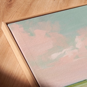 May include: A framed canvas artwork with a pastel landscape design. The painting uses soft pink, light blue, and green hues, set within a natural wood frame. The artwork creates a calm, airy feel, ideal for interior decoration.