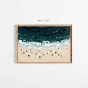 Könnte beinhalten: Ein Ölgemälde zeigt eine Strandszene mit dunkel türkisblauen Wellen, die auf einen sandigen Strand treffen. Kleine Figuren von Menschen sind über den Strand verstreut. Das Kunstwerk ist in einem hellen Holzrahmen gerahmt, mit dem Text "VINTAGE GLOW PRINTS" oben.