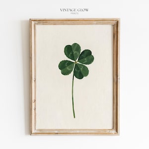 May include: A framed wall art print featuring a centred illustration of a four-leaf clover with deep green leaves and a thin stem on a textured off-white background. The text VINTAGE GLOW PRINTS is printed in black at the top centre of the frame.