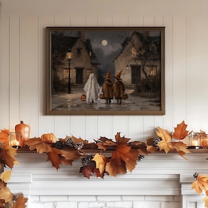 Op de afbeelding: Een ingelijste kunstdruk met een nachtelijke sc&egrave;ne van drie figuren die over een kasseienstraat tussen twee huizen lopen. E&eacute;n figuur draagt een wit spookkostuum, twee dragen oranje heksenhoeden en capes, en ze dragen allemaal pompoenen.