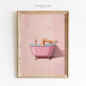 May include: A framed art print titled VINTAGE GLOW PRINTS featuring a pink clawfoot bathtub with a light blue rim. A pair of legs and a hand holding a martini glass with a lime garnish emerge from the tub against a solid light pink background.