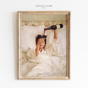 Può includere: Stampa incorniciata raffigurante una persona in accappatoio bianco che versa vino rosso in un bicchiere mentre è a letto. La stampa ha un'estetica vintage, con il testo "VINTAGE GLOW PRINTS" in alto.