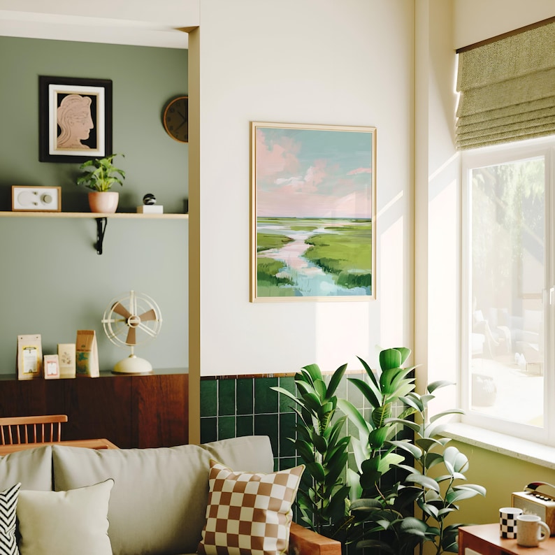 May include: A framed landscape painting shows a marsh scene with a river winding through green fields under a pink and blue sky. The artwork is displayed in a living room with a sofa, plants, and decorative items.