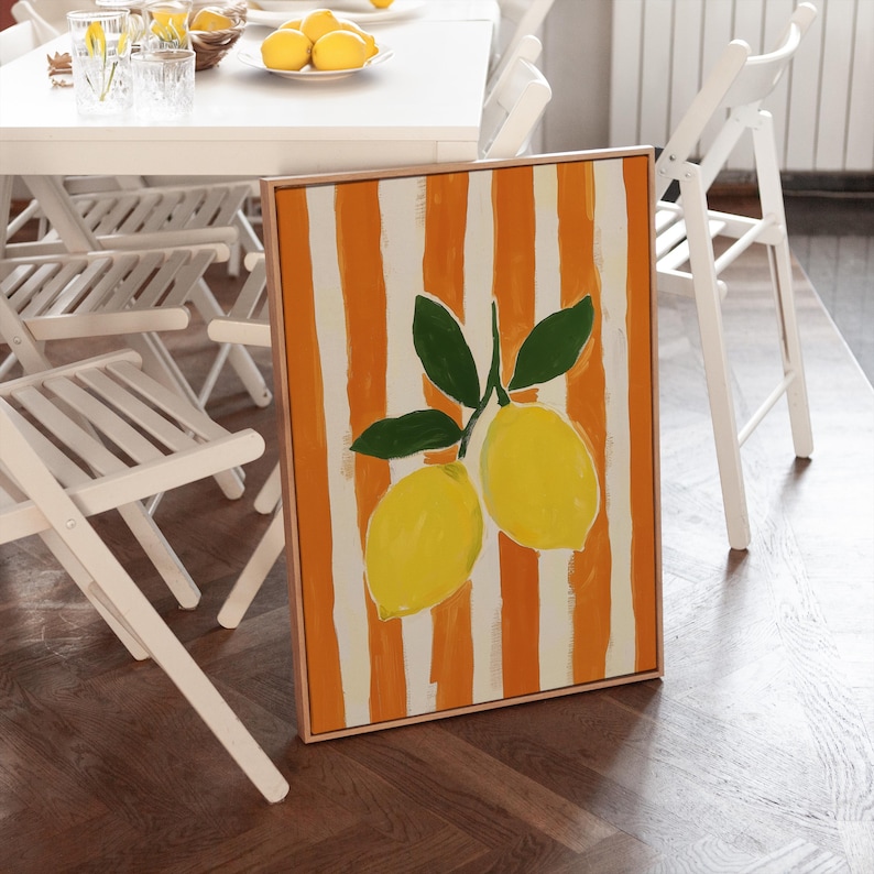 May include: A framed painting featuring two yellow lemons with green leaves against a background of orange and white vertical stripes. The artwork is displayed on a wooden floor, with white chairs and a table in the background.