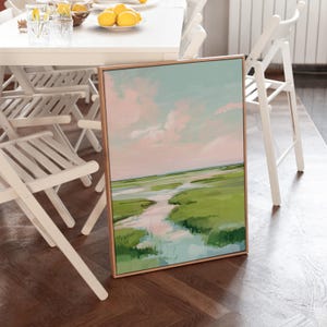 May include: A framed landscape painting depicting a marsh scene. The artwork shows a light blue sky with pink clouds, a green marsh, and a waterway. The frame is a light brown colour.