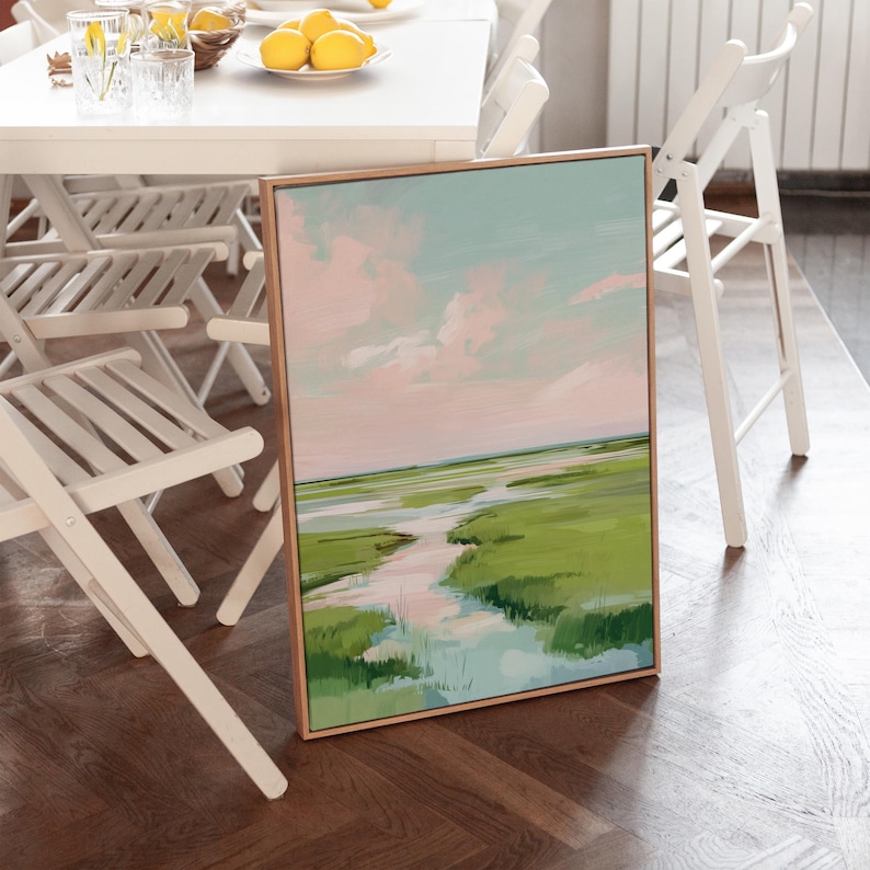 May include: A framed landscape painting depicting a marsh scene. The artwork shows a light blue sky with pink clouds, a green marsh, and a waterway. The frame is a light brown colour.