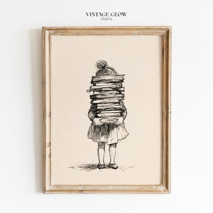 May include: A framed wall art print featuring a black ink line drawing of a person seen from behind, holding a tall, precarious stack of books. The person has hair tied in a bun and wears a dress. The text VINTAGE GLOW PRINTS appears at the top.