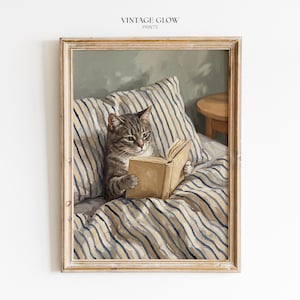 Könnte beinhalten: Ein gerahmter Kunstdruck mit dem Titel VINTAGE GLOW PRINTS zeigt eine getigerte Katze, die aufrecht in einem Bett mit blau-weiß gestreifter Bettwäsche sitzt und ein offenes Buch hält. Die Katze lehnt an zwei gestreiften Kissen neben einem Holztisch.