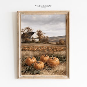 Op de afbeelding: Een ingelijste kunstdruk getiteld VINTAGE GLOW PRINTS met een olieverfschilderij van een landelijk landschap. De scène toont een veld met oranje pompoenen op de voorgrond, een witte boerderij, herfstbomen en heuvels onder een bewolkte lucht.