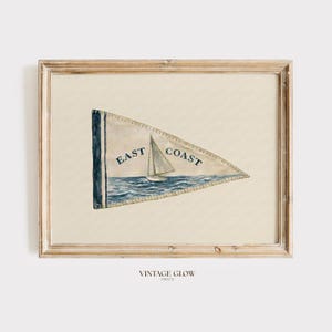 May include: A framed print featuring a triangular pennant flag with a cream background. The flag displays a blue sailboat on water with the text EAST COAST in dark blue lettering. The frame is made of light-colored wood with a natural grain finish.