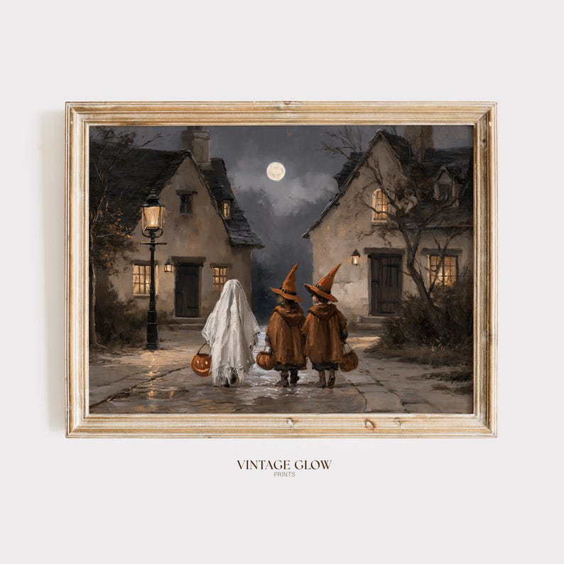 Op de afbeelding: Een ingelijste kunstdruk getiteld VINTAGE GLOW PRINTS met een nachtelijke sc&egrave;ne van drie figuren in Halloween-kostuums die over een geplaveide straat tussen twee huizen lopen onder een volle maan, waaronder een geest en twee heksen met pompoenen.