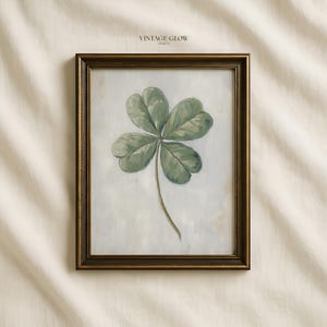 May include: A framed print featuring a detailed painting of a green shamrock with three leaves. The artwork is set against a soft, off-white background and framed in a dark, ornate gold frame. The text "VINTAGE GLOW PRINTS" is at the top.