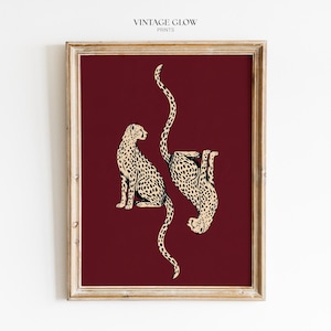 Peut inclure: Une impression d'art encadrée représentant deux guépards couleur crème avec des taches noires sur un fond bordeaux profond. Un guépard est assis droit tandis que l'autre est positionné à l'envers. Le texte VINTAGE GLOW PRINTS apparaît en haut.