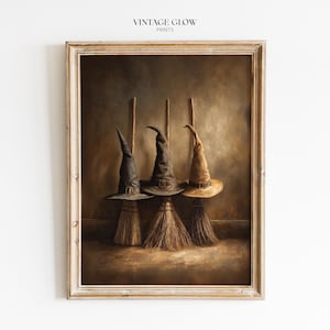 Op de afbeelding: Een ingelijste kunstdruk getiteld VINTAGE GLOW PRINTS met drie heksenhoeden in zwart, donkergrijs en beige, elk rustend op een houten bezemsteel. De hoeden hebben puntige kronen en gespen, geplaatst tegen een donkere, getextureerde bruine achtergrond.