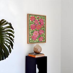 以下が含まれることがあります： 淡い黄色の背景にピンクのピオニーの花と緑の葉が描かれた鮮やかなイラストの長方形の額入りウォールアート。濃い緑色の縁取りが施され、明るい木製のフレームに入れられて白い壁に飾られています。