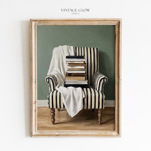 May include: A framed print featuring a vintage-style interior scene. A striped armchair holds a stack of books and a white blanket. The background is a muted green, and the frame has a gold finish. The text "VINTAGE GLOW PRINTS" is at the top.