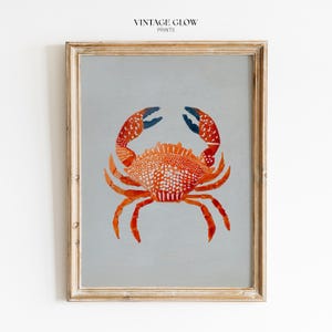 May include: A framed wall art print featuring a vibrant orange crab with white speckled details and dark blue claw tips centered on a solid light gray background. The text VINTAGE GLOW PRINTS is printed in small black letters at the top center.