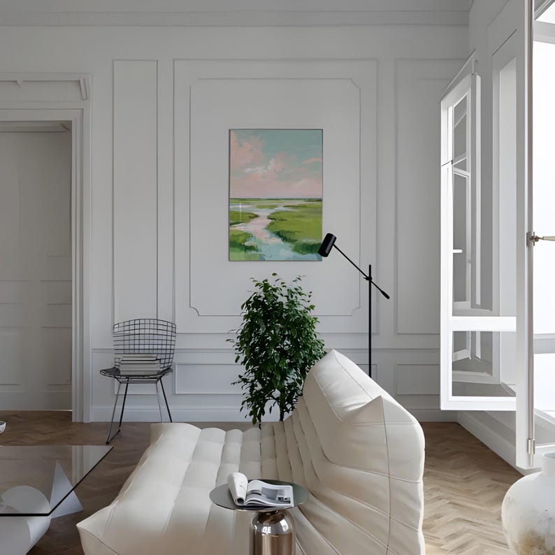 May include: A modern living room featuring a white sofa, a black floor lamp, and a painting depicting a marsh landscape. The artwork shows a river winding through green marshland beneath a pink and blue sky. A small table holds a book and a plant.