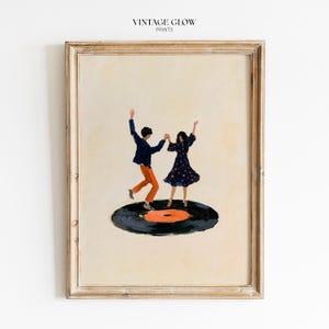 Op de afbeelding: Een ingelijste kunstdruk met de tekst VINTAGE GLOW PRINTS toont twee dansende figuren op een zwarte vinylplaat. Eén figuur draagt een donkerblauw jasje en een oranje broek, de andere een donkerblauwe jurk met oranje stippen op een crèmekleurige achtergrond.