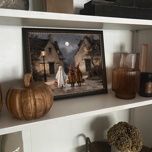 Op de afbeelding: Een ingelijste rechthoekige print met een nachtelijke sc&egrave;ne van een geest en twee figuren met puntmutsen die over een geplaveide straat tussen twee huizen lopen onder een volle maan, verlicht door een lantaarnpaal en warme ramen.