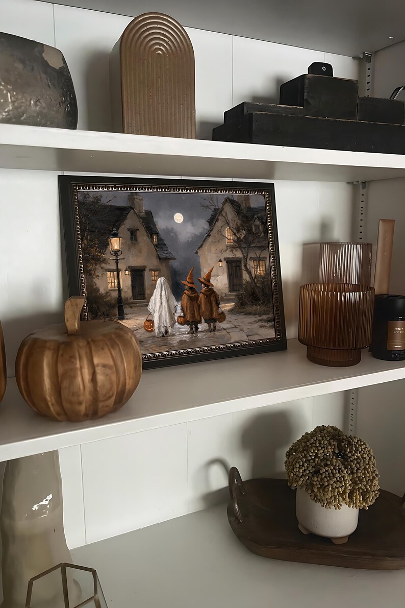 Op de afbeelding: Een ingelijste rechthoekige print met een nachtelijke sc&egrave;ne van een geest en twee figuren met puntmutsen die over een geplaveide straat tussen twee huizen lopen onder een volle maan, verlicht door een lantaarnpaal en warme ramen.