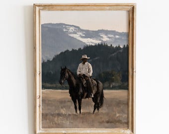 Op de afbeelding: Een ingelijste kunstdruk getiteld VINTAGE GLOW PRINTS met een schilderij van een persoon in een licht overhemd en cowboyhoed op een donker paard in een veld. Op de achtergrond zijn donkere beboste heuvels en besneeuwde bergen te zien.