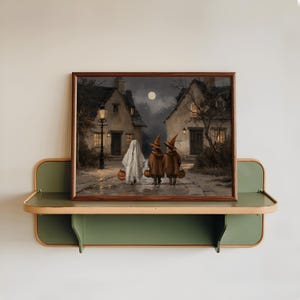 Op de afbeelding: Een ingelijste rechthoekige wandkunstprint met een nachtelijke sc&egrave;ne van een geest en twee figuren met puntmutsen die onder een volle maan over een kasseienstraat tussen huizen lopen, elk met een kleine oranje emmer in de vorm van een pompoen.