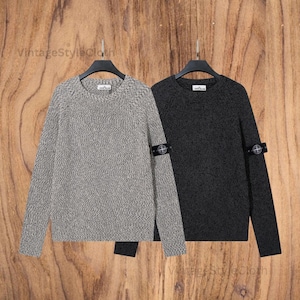 Può includere: Due maglioni girocollo, uno grigio chiaro e uno grigio scuro, sono appesi a grucce nere. Ogni maglione ha una piccola toppa sulla manica sinistra. I maglioni sono esposti su uno sfondo di legno. Il testo "VintageStyleCloth" è visibile.