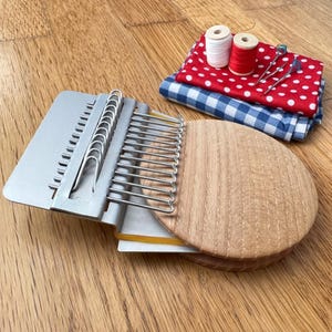 May include: A silver metal weaving tool with a wooden base is open, displaying metal loops. Two spools of thread, red and white, sit next to red polka dot and blue gingham fabric. Sewing pins are also visible.