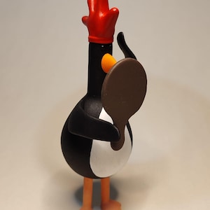 Puede incluir: Una figurita de pingüino blanco y negro con un sombrero rojo y una paleta marrón en su mano. El pingüino está de pie sobre una superficie blanca.