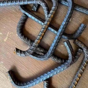 May include: A collection of bent, dark gray steel rebar pieces with a textured surface. The rebar has a curved, J-shaped design, and some pieces show signs of rust. The pieces are scattered on a wooden surface.