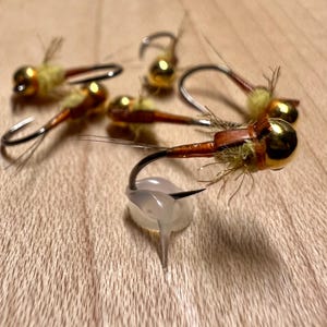 May include: Close-up of five gold and brown fly fishing lures with yellow and brown feathers. The lures are tied on hooks and have a gold bead at the head.