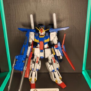 May include: A detailed model of the MSZ-010 ZZ Gundam, a mecha robot, in a display. The robot is primarily white, blue, and red, with a detailed design. The model includes a beam saber and missile launcher. The text on the display reads "MSZ-010 ZZ GUNDAM".