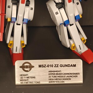May include: A detailed image of a white, blue, red, and yellow MSZ-010 ZZ Gundam model. The model has articulated limbs and is standing on a surface. A metal plate displays the model's name, height (22.11 meters), and weight (68.4 metric tons).