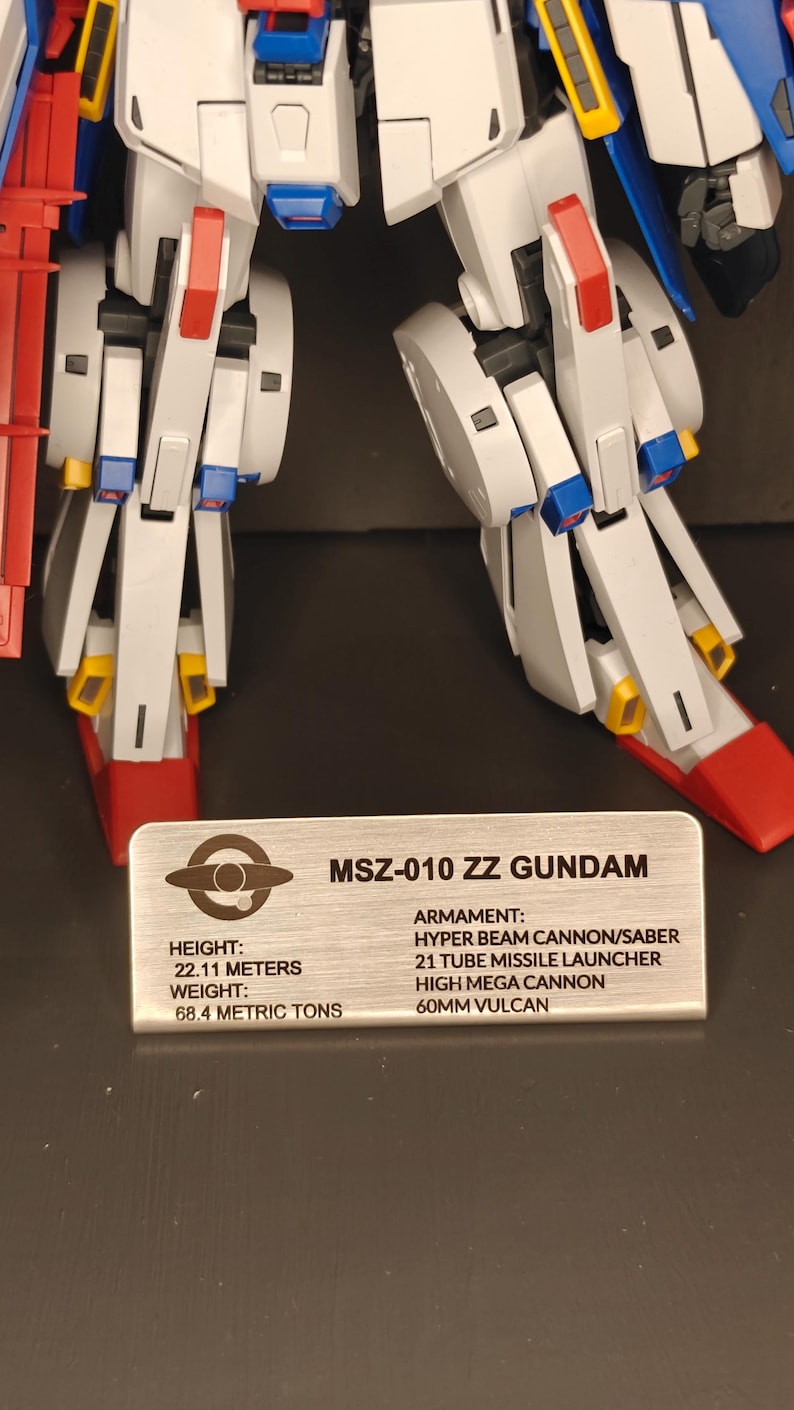 May include: A detailed image of a white, blue, red, and yellow MSZ-010 ZZ Gundam model. The model has articulated limbs and is standing on a surface. A metal plate displays the model's name, height (22.11 meters), and weight (68.4 metric tons).