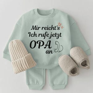 Könnte beinhalten: Ein hellgrünes Baby-Outfit-Set mit passender Mütze und Hausschuhen. Sweatshirt und Hose tragen den Text "Mir reicht's Ich rufe jetzt OPA an" in Schwarz. Die Mütze ist beige.