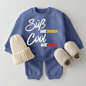 Könnte beinhalten: Ein blaues Kinder-Outfit-Set mit Sweatshirt, Hose, Mütze und Hausschuhen. Das Sweatshirt hat die Worte "Süß Cool" in weißer Schrift, mit "wie Mama" in Gelb und "wie Papa" in Rot. Mütze und Hausschuhe sind beige.