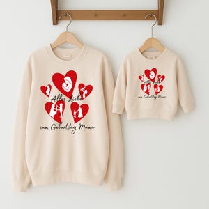 Könnte beinhalten: Zwei beige Sweatshirts, eines für Erwachsene und eines für Kinder, hängen an Holzbügeln. Jedes Sweatshirt hat ein Design aus roten Herzen mit Silhouetten und dem deutschen Text "Alles Liebe zum Geburtstag Mama".