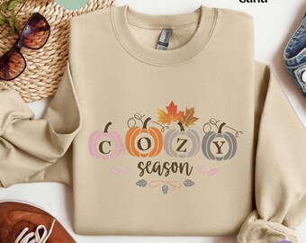 May include: Sand-colored sweatshirt with the word "COZY" spelled out with pumpkin graphics. The word "season" is written below the pumpkins. The sweatshirt has a crew neck and long sleeves.