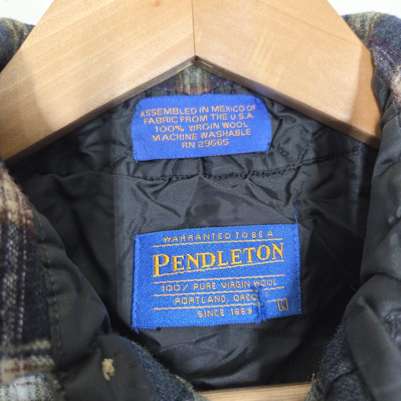 May include: Close-up of a Pendleton jacket label. The blue label reads "WARRANTED TO BE A PENDLETON 100% PURE VIRGIN WOOL PORTLAND, OREG. SINCE 1869." Another blue label states "ASSEMBLED IN MEXICO OF FABRIC FROM THE U.S.A. 100% VIRGIN WOOL MACHINE WASHABLE RN 29565."