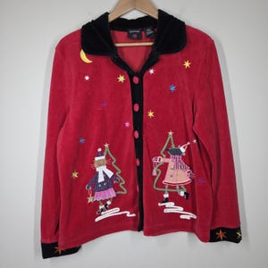 May include: Red button-up jacket with black velvet collar and trim. Features embroidered Christmas-themed designs, including a cat and a mouse, Christmas trees, stars, and a crescent moon. The jacket has long sleeves and a relaxed fit.
